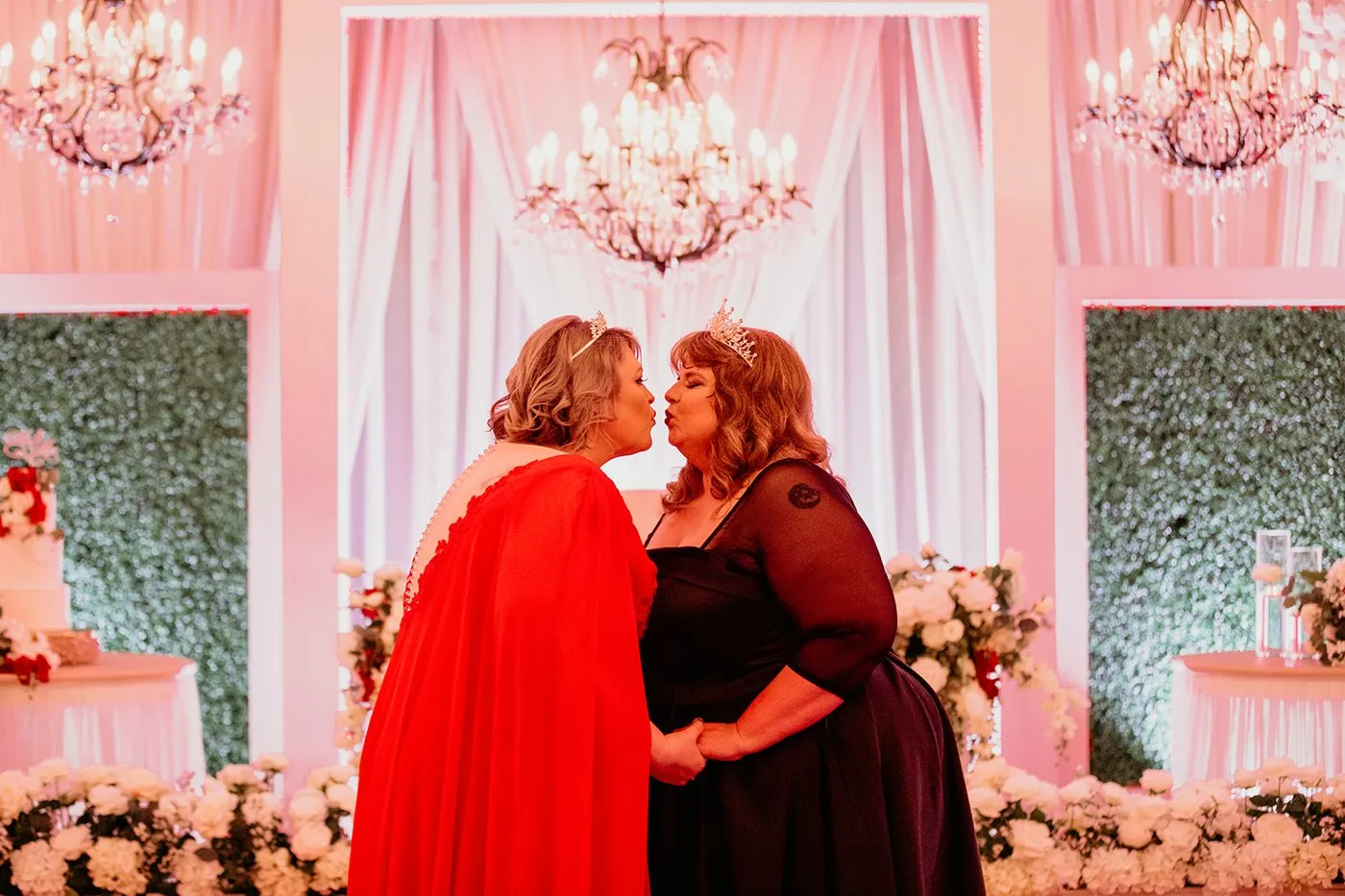 Two brides in tiaras sharing a kiss beneath a chandelier at their reception
