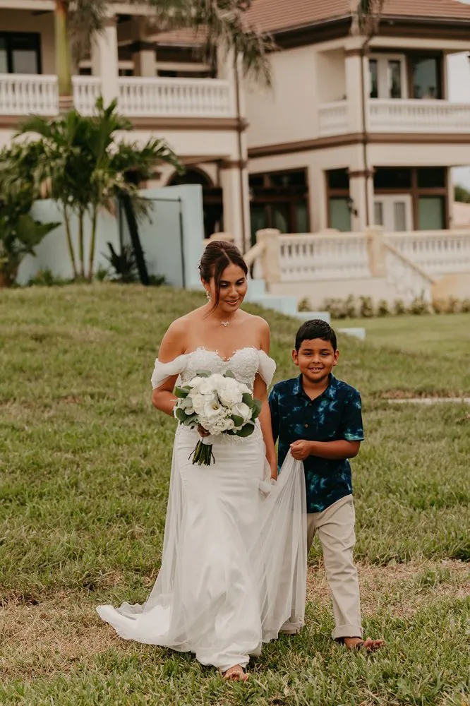 Camila caminando mientras un niño le lleva la cola del vestido frente a la mansión