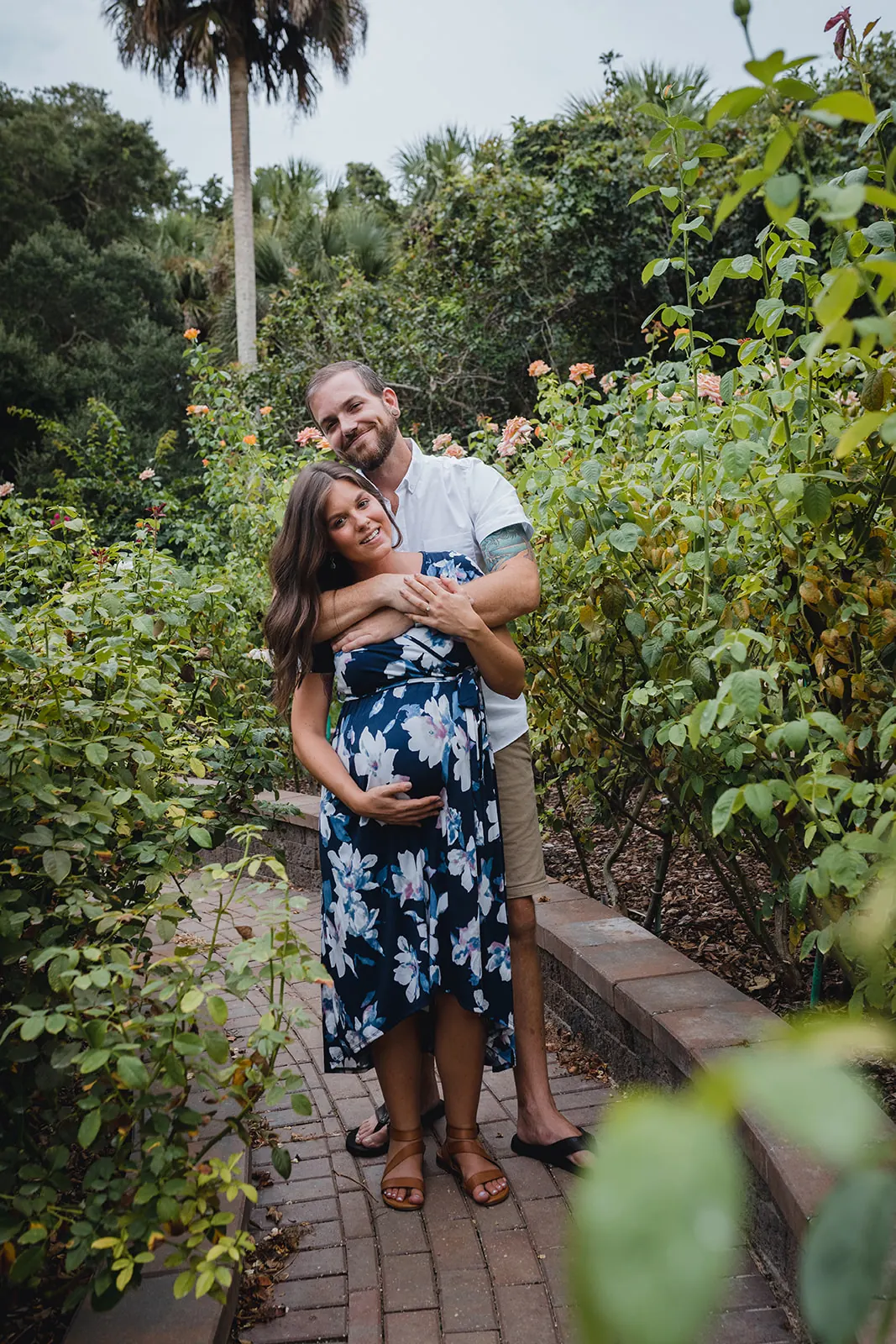 Jordan embracing Christine from behind in a rose garden during their maternity session