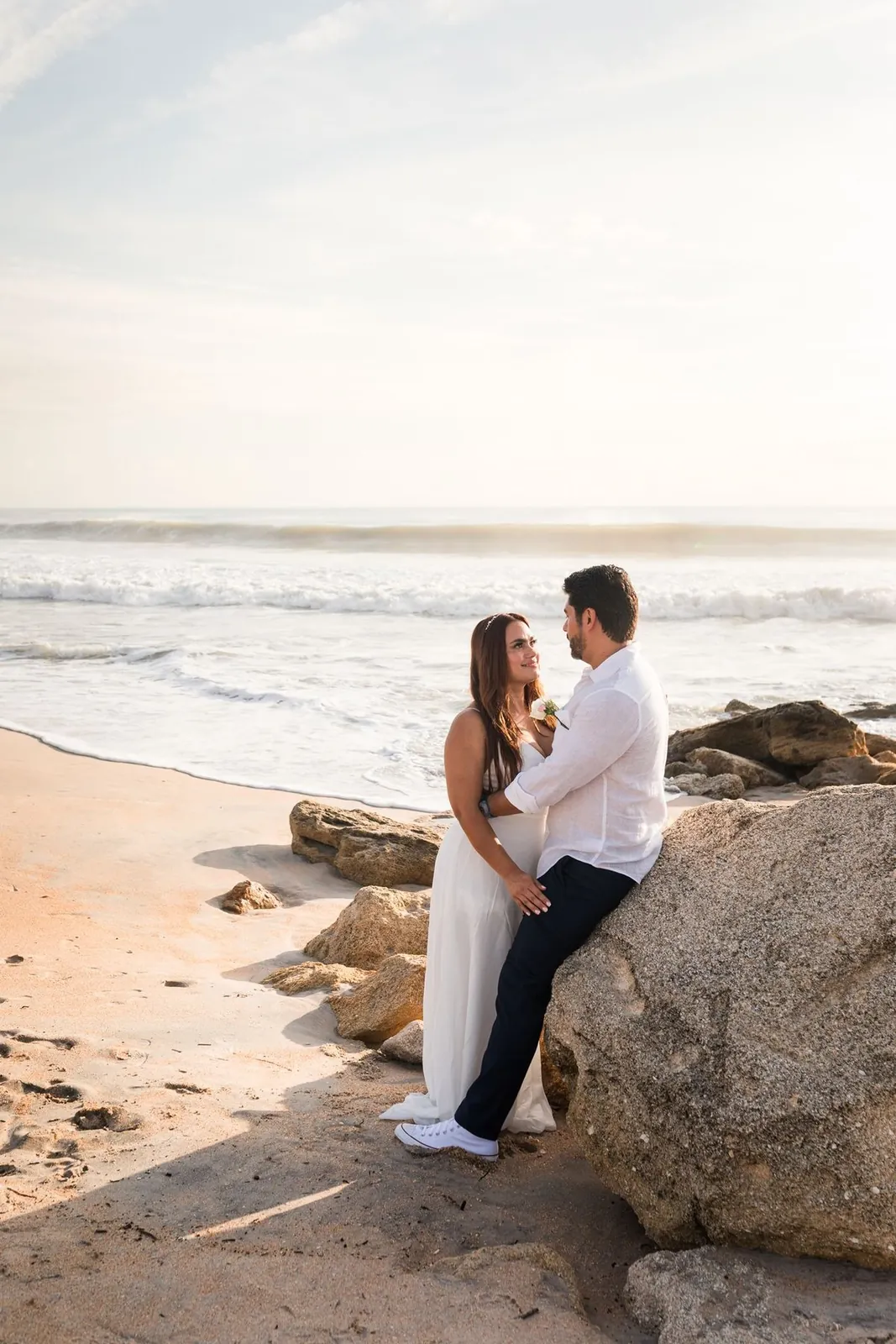 Bride and groom on the beach at golden hour, leaning against the rocks