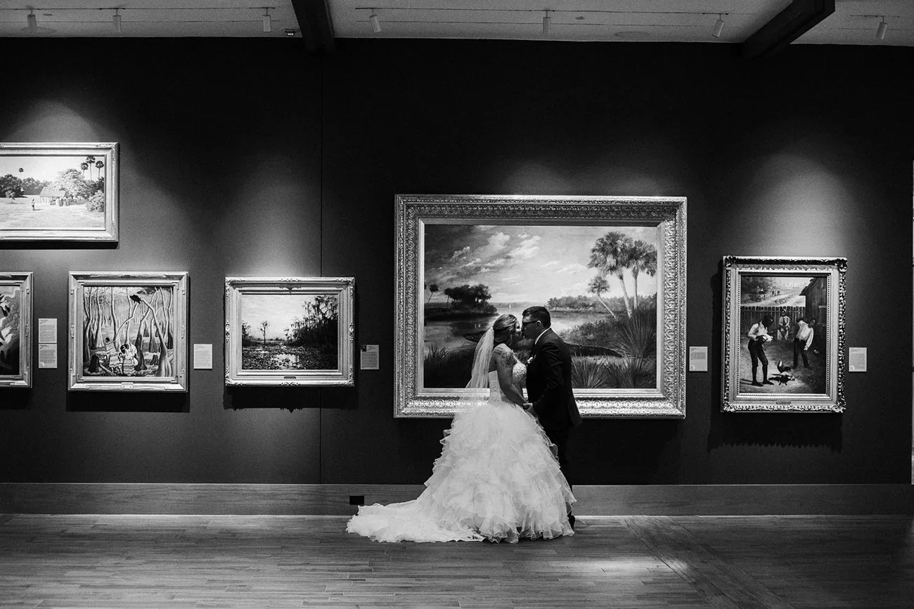 Haley and Matt kissing in black and white before a large framed Florida landscape painting