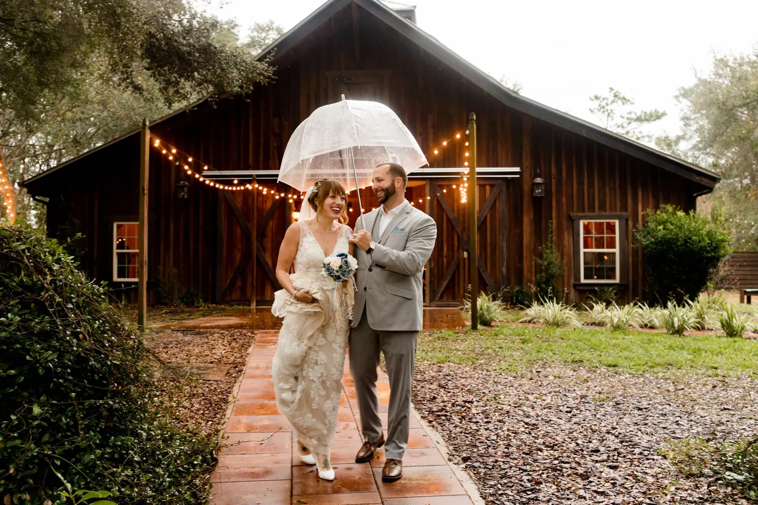 A bride and groom walking under an umbrella past a rustic barn in the rain