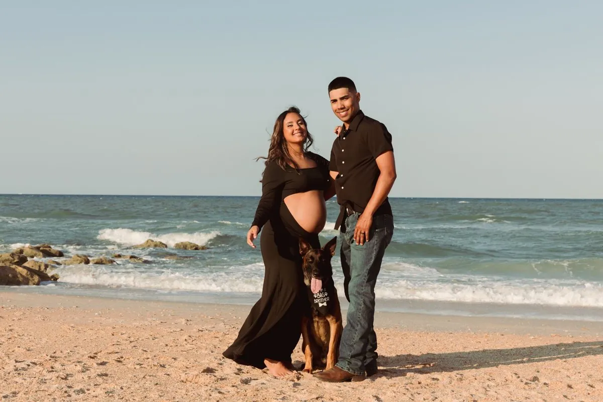 An expectant couple on the beach with their German shepherd wearing a Big Brother bandana