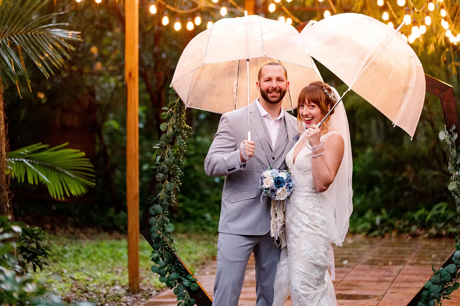 A bride and groom laughing under clear umbrellas at their rainy garden wedding