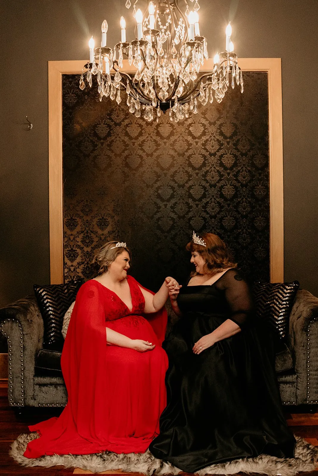 Two brides in red and black gowns holding hands on a velvet sofa