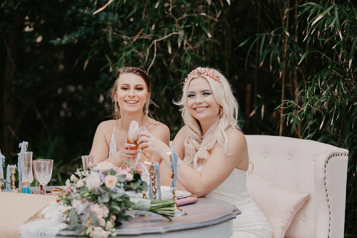 Two brides toasting with drinks at their sweetheart table surrounded by greenery and pastel florals