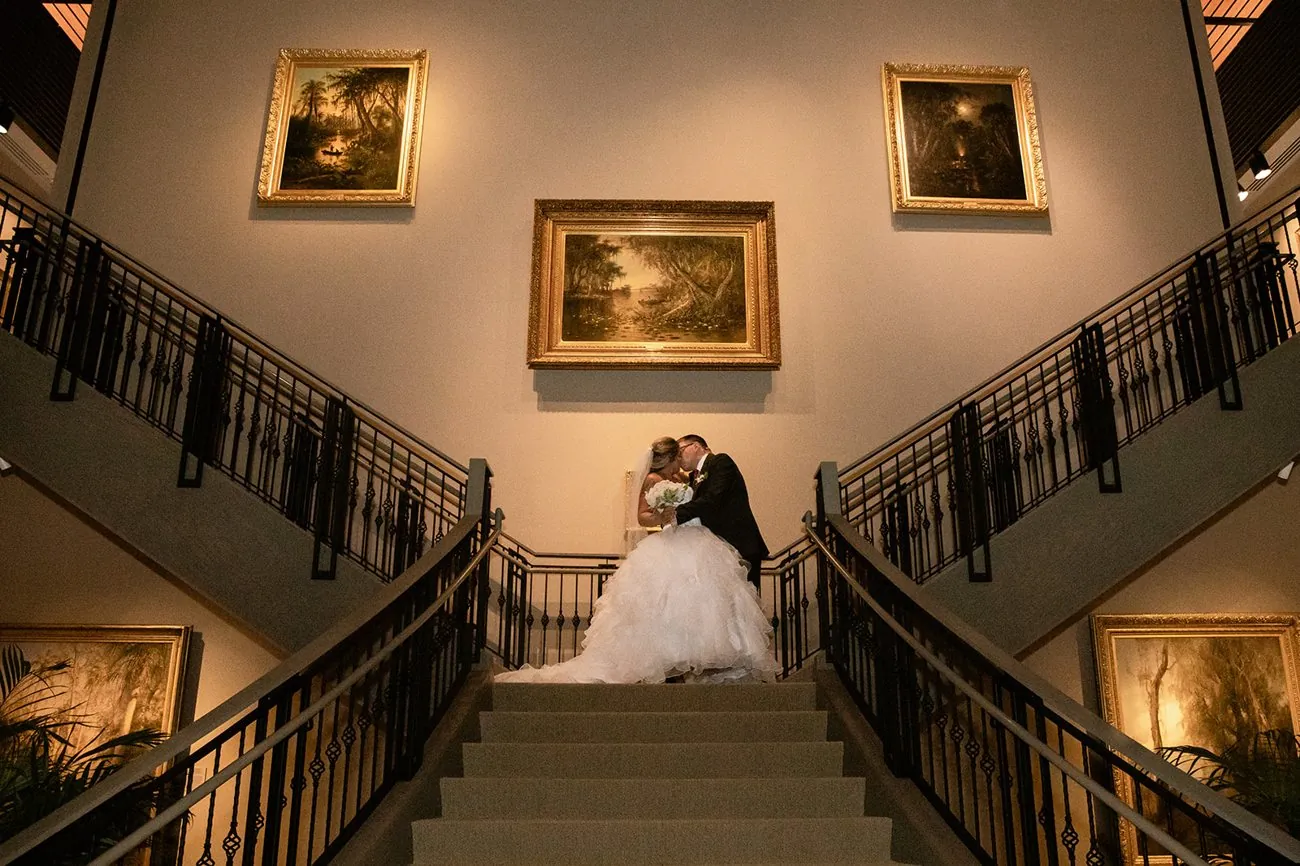 Haley and Matt kissing on the grand staircase beneath gold-framed Florida paintings at the Cici and Hyatt Brown Museum