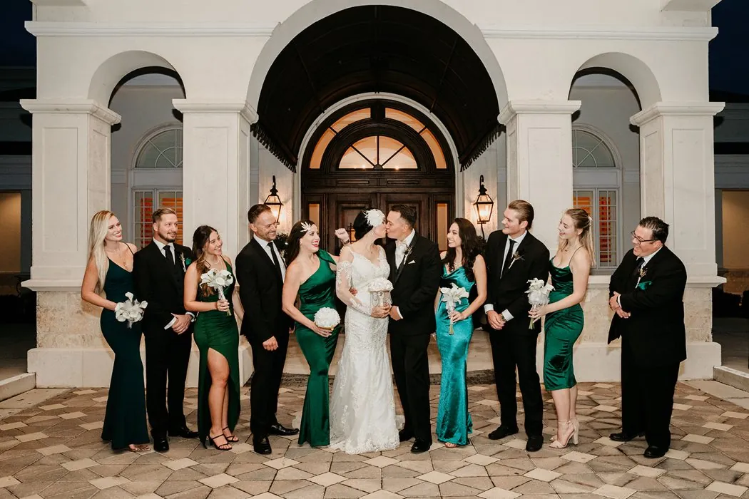 The wedding party in emerald and black tuxedos as the couple kisses at a Gatsby wedding
