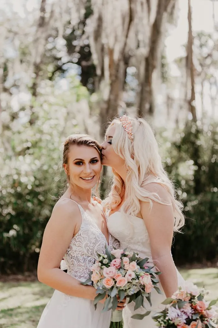 Two brides holding pastel bouquets, one kissing the other on the temple