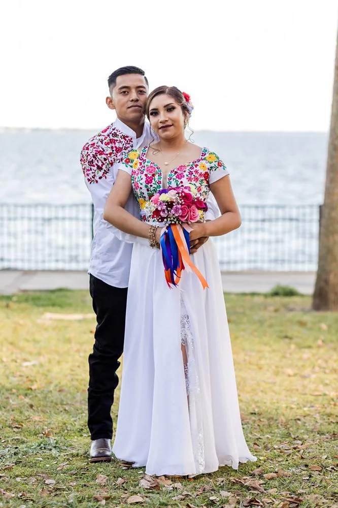 Nati y Juan abrazados con trajes bordados tradicionales mexicanos junto al agua